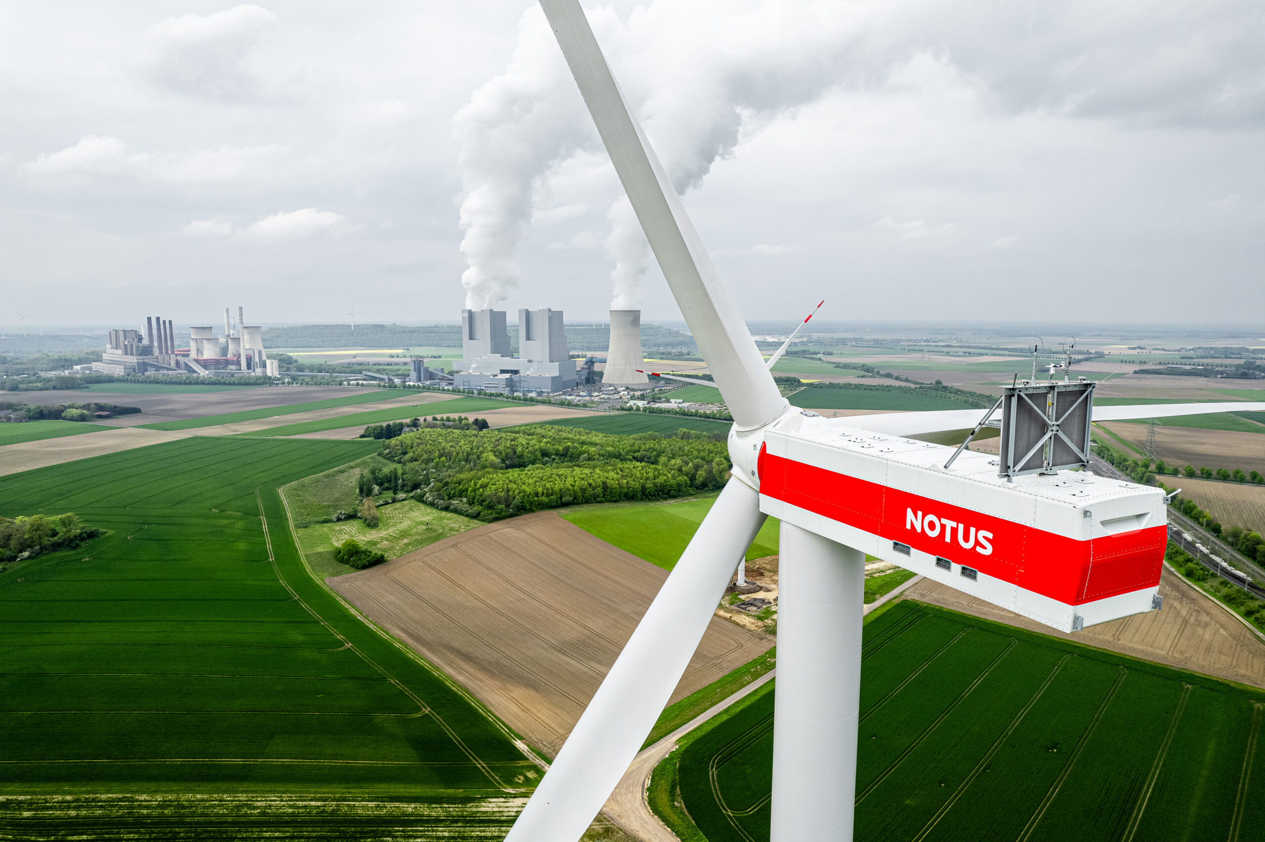 Im Vordergrund das Maschinenhaus eines Windrads mit der Aufschrift Notus. Im Hintergrund grüne Feldern und Kraftwerke.