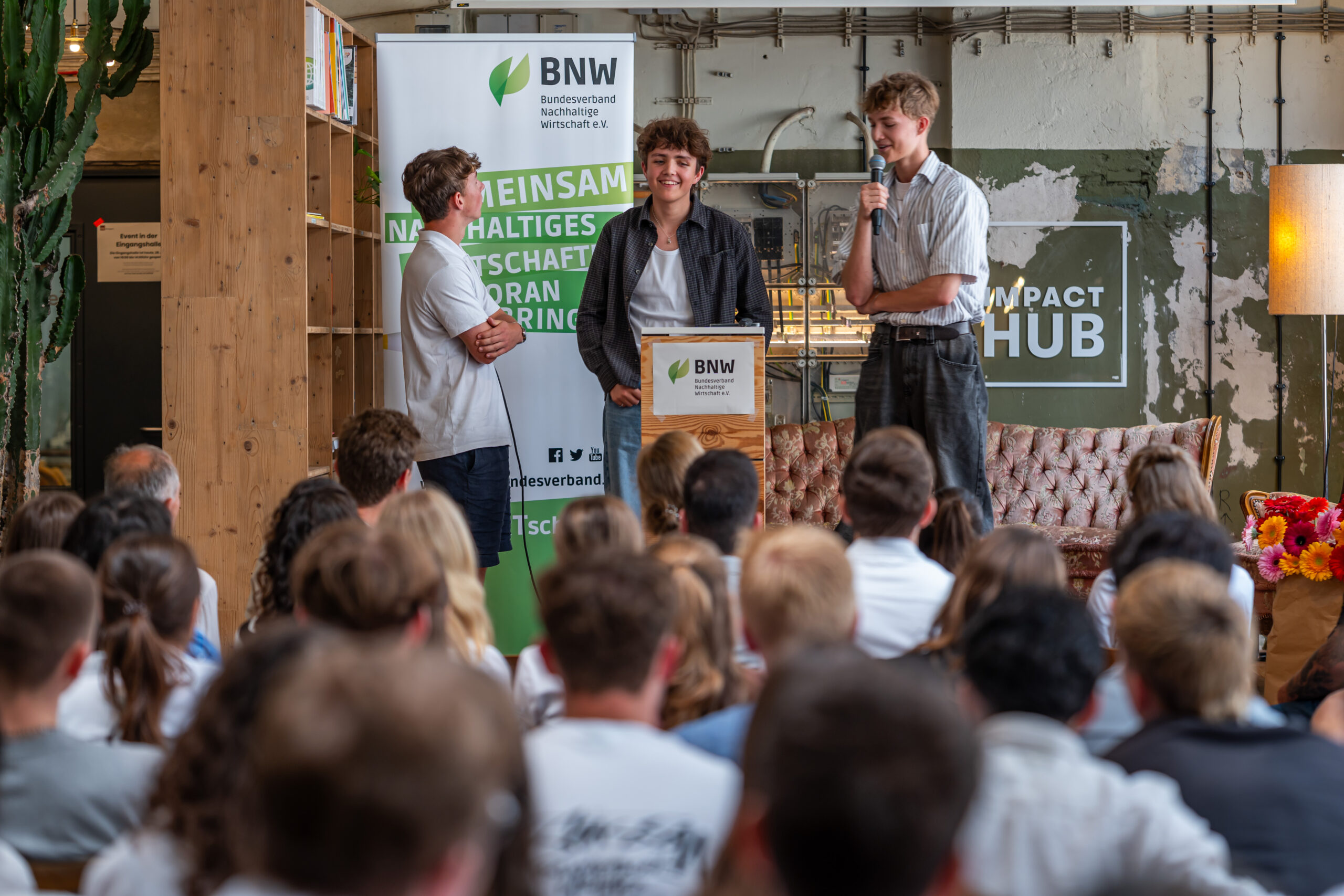 Drei jugendliche Kinder stehen auf einem Podium vor einem Publikum und stellen ihr Projekt vor. Im Hintergrund ist ein Ausfsteller des BNW zu sehen.